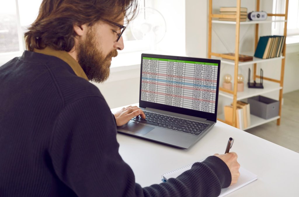 A bearded man sitting at office desk with notebook PC, working with a spreadsheet and writing down information in notepad