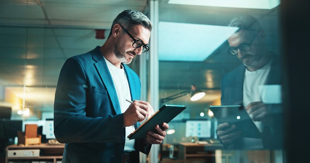 A man wearing glasses and a blue blazer stands in a modern office, writing on a tablet with a stylus