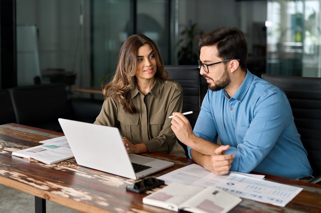 Two busy colleagues working together talking using laptop in office.